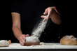 Making bread. Hands of flour splatter and fly onto the white table covered with flour from his hands. The cook sprinkles white flour dust with a man's hand against a black background. Space for text.