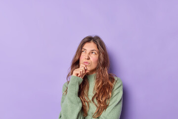 Wall Mural - Thoughtful young Caucasian woman keeps hand on chin frowns face thinks above something concentrated above wears casual jumper isolated over purple background copy space for your advertising content