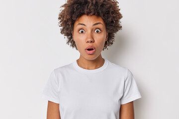 Wall Mural - Photo of startled curly haired woman holds breath has eyes popped out cannot believe in shocking news wears casual t shirt isolated over white background stands anxious. Human reactions concept