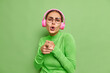 © WHstudio Leushin N - Glad playful woman sings song indicates at you enjoys favorite music in pink headphones enjoys moment wears transparent glasses and turtleneck poses against green background. Monochrome shot