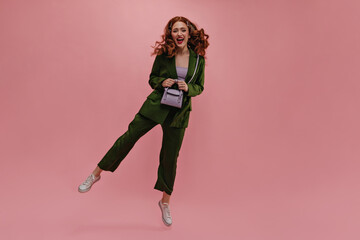 Full-length cheerful pretty caucasian young girl bounces while in photo studio. Wavy red hair flying away from european woman. Dressed in green suit and bag in hands. Energy concept of human life