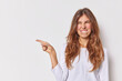 © WHstudio Leushin N - Cheerful positive woman with long hair has happy expression bites lips points index finger at blank space shows logo or banner stands against white background demonstrates company advertisement