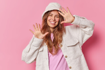 Wall Mural - Horizontal shot of good looking young woman has playful expression raises palms smiles joyfully entertains you wearsfur jacket and hat poses against pink background. Positive emotions concept