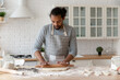 © fizkes - Focused young African American man baking cookies in home kitchen. Food blogger, baker, chef in apron rolling dough for biscuit, bread, cake, cooking bakery dessert at flour table with ingredients.