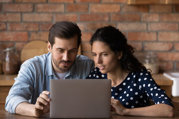 Important information. Focused millennial husband wife sit by laptop at kitchen table do shopping online. Thoughtful young couple choose bank at web read mortgage loan terms conditions on pc screen