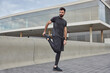 © WHstudio Leushin N - Full length shot of active muscular adult man raises legs warms up before workout wears black t shirt shorts and sneakers trains outdoors in urban setting. Healthy lifestyle and sport concept