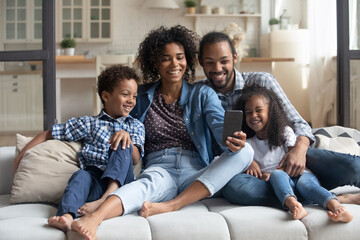 Wall Mural - Happy African couple of parents and laughing kids resting on couch holding smartphone, taking selfie on cellphone, making video call, looking at mobile phone screen, smiling, giggling. Family leisure