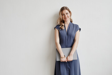 Wall Mural - Minimal portrait of beautiful blonde woman holding laptop and smiling at camera while standing by white wall, copy space