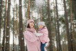 © sofiko14 - Young mother holding cute little daughter, in a winter forest for a walk. Pretty mom with child on hands, wearing pink coats throwing snow up and laughing. Family in winter forest