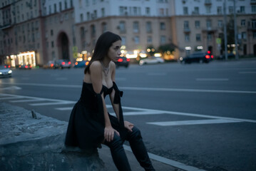 beautiful brunette girl in a black dress is sitting by the high road in the evening. Attractive professional languid look traffic cars