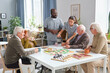 © pressmaster - Nurse and doctor pouring hot tea for senior people while they sitting at the table and playing board games in nursing home