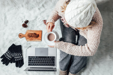 Wall Mural - A girl holding a cup of hot chocolate while working with laptop at home on winter