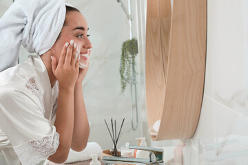 Poster - Beautiful young woman applying cleansing foam onto face near mirror in bathroom, space for text. Skin care cosmetic