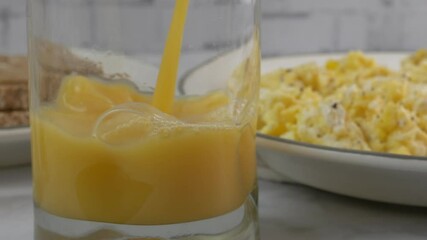 Wall Mural - Pouring orange juice into a glass by a plate of scrambled eggs slow motion
