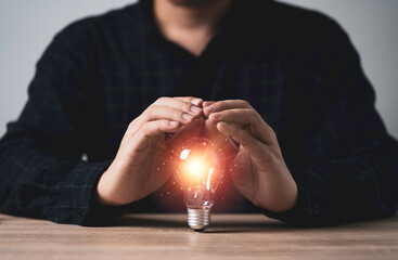 Poster - businessman hand protecting glowing light bulb with orange light ray on wooden table for inspiration