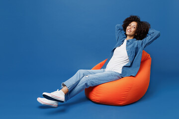 Wall Mural - Full body young smiling happy black woman in casual clothes shirt white t-shirt sit in bag chair hold hands behind neck relax isolated on plain dark blue background studio. People lifestyle concept.