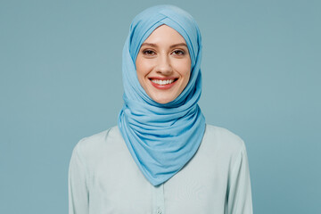Young smiling calm confident arabian asian muslim woman in abaya hijab clothes look camera isolated on plain blue color background studio portrait. People uae middle eastern islam religious concept.