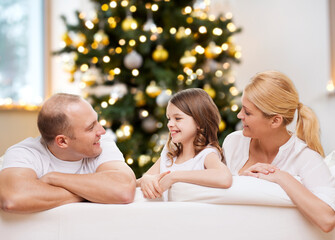 Poster - family, winter holidays and technology concept - portrait of happy mother, father and daughter sitting on sofa at home over christmas tree lights background