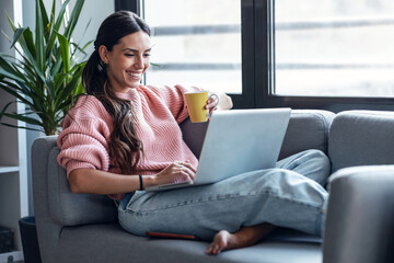 Wall Mural - Cute woman having videocall with friends on laptop sitting on the couch at home.