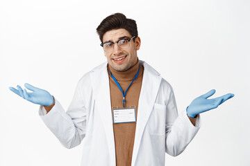 Wall Mural - Image of doctor in glasses and medical uniform shrugging shoulders, smiling confused, dont know, standing over white background