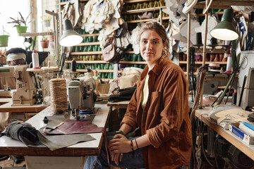 Portrait of young tailor looking at camera while sitting at her workplace with sewing machine in the factory