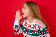 © Roquillo - brunette kid girl in knitted sweater christmas over red background look empty space holding hand near her face and screaming or calling someone.