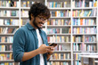 © Stock 4 You - Side view portrait of happy young Indian student chatting by mobile phone in university library. Teen guy wearing denim shirt and eyeglasses using social media application browsing on smartphone