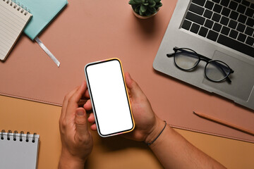Poster - Top view image of a hand using a white blank screen smartphone over the working desk surrounded by a potted plant, computer laptop, notebook, pencil and eyeglasses.