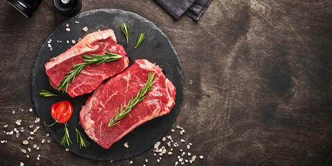 Wall Mural - Two raw steak New York  with rosemary and spices on a piece of parchment paper on old dark stone background. Top view. Mock up.