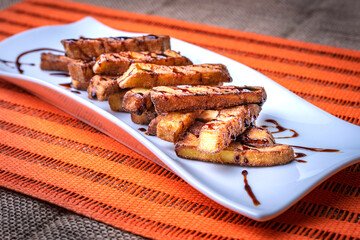 Wall Mural - Presentation in rectangular plate of aubergines cut into strips and fried, with sugar cane honey scattered on top, on an orange background.