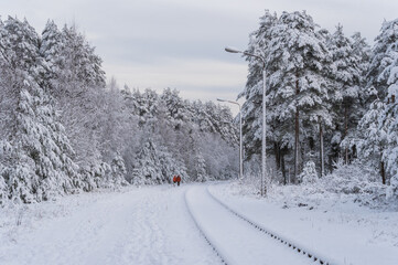 Two unrecognizable people in red jackets walking by the abandoned railway rails in snowy white forest
