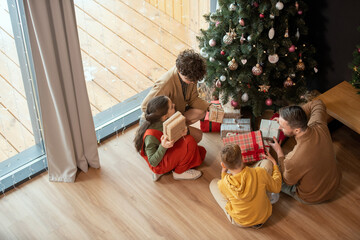 Sticker - Above view of family with two children sitting on floor near Christmas tree and shaking gift boxes while guessing what Is inside of it