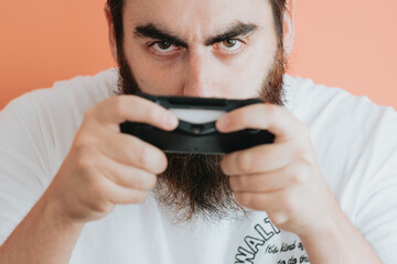 Young hipster bearded man concentrated holding a console pad, video games concept, copy space, soft orange removable background, minimal basic, ad concept deal, white shirt space