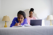 Teenage Girl Lying On Bed And Writing In Notebook, Mother Using Laptop In Background