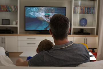 Poster - Rear view of father and son sitting at home together watching swimming competition on tv