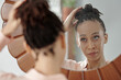© DragonImages - Beautiful young Black woman looking at mirror and making hair bun