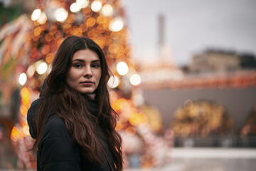 Sticker - A beautiful and young girl looks into the camera. Christmas city decoration blurred in the background. Cute brunette faced to the camera in the downtown before New Year party