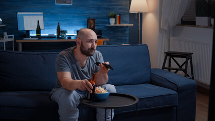Wall Mural - Bored man changing tv channels sitting on cozy couch drinking beer, home alone late at night after work. Person relaxing watching television holding remote control searching a comedy movie.