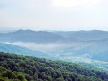 Landscape Of A Mountain Village Surrounded By Green Waves Of Beech Forests Under The Fog Hanging In The Air.