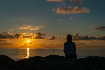 Young woman in a white shirt and jeans is sitting on huge rocks and looking at a beautiful, mesmerizing sunset over the sea, photo from the back, copy space