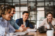 © Maskot - Smiling businessman looking away while sitting amidst cheerful colleagues at conference table