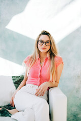 Wall Mural - Portrait of caucasian woman in eye glasses sitting on a sofa on green pastel background in studio.