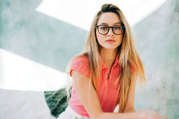 Wall Mural - Portrait of caucasian woman in eye glasses sitting on a sofa on green pastel background in studio.