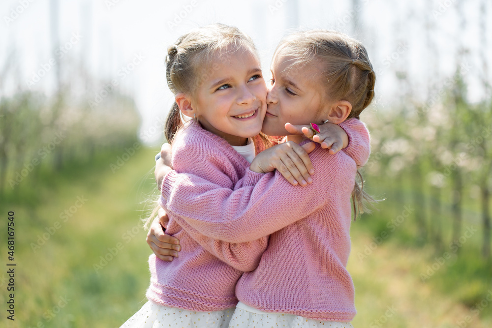 Happy Twin Sisters Hug Against The Background Of A Green Blooming Apple happy-twin-sisters-hug-against-the-background-of-a-green-blooming-apple