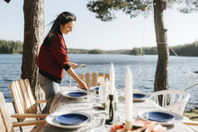 Smiling Young Woman Setting Table By Lake On Sunny Day