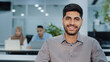 © Yuliia - Confident happy arab entrepreneur young male top manager portrait. Smiling company owner, startup businessman, leader, successful employee looking at camera. His subordinates working on background