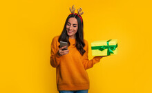 Close Up Photo Of Happy Stylish Woman In Sweater And Christmas Hat With Present Box In Hand Is Using Smart Phone Isolated On Yellow Background
