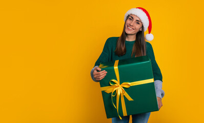 Wall Mural - Beautiful happy stylish woman in christmas Santa hat with present box in hands is posing isolated on yellow background