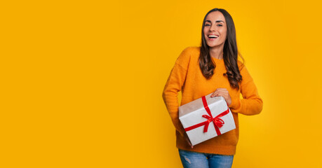 Attractive excited smiling young woman holds beautiful gift box in hands and looks on the camera isolated on yellow background