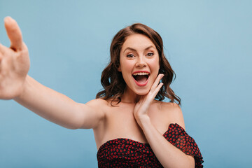 Poster - Close-up of happy brown-haired woman with loose hair makes selfie on blue background. Holding her face with open mouth, girl looks at camera dressed in black outfit with bare shoulders.